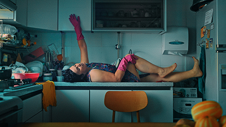 Filmstill aus "Becoming Kim": Ansicht einer Person, die ausgestreckt in der Küche auf der Küchentheke liegt.